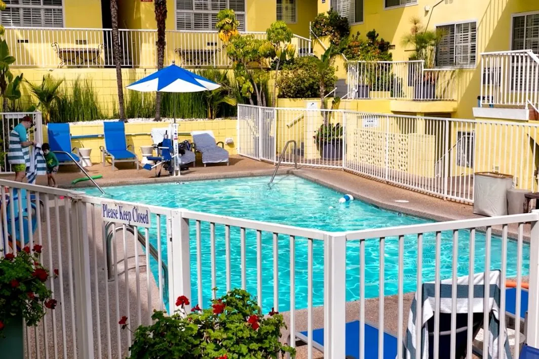 A fenced pool area with blue lounge chairs and a yellow building in the background.