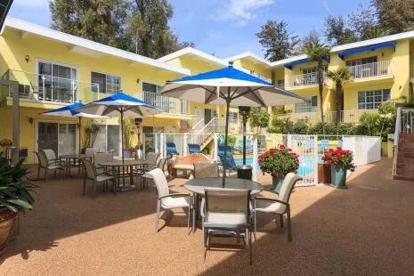 A sunny outdoor pool area with yellow buildings, blue and white umbrellas, and flowering plants.