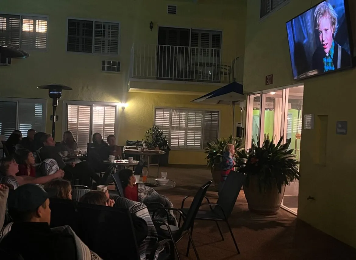 A group of people sitting in an outdoor courtyard watching a movie on a large screen.