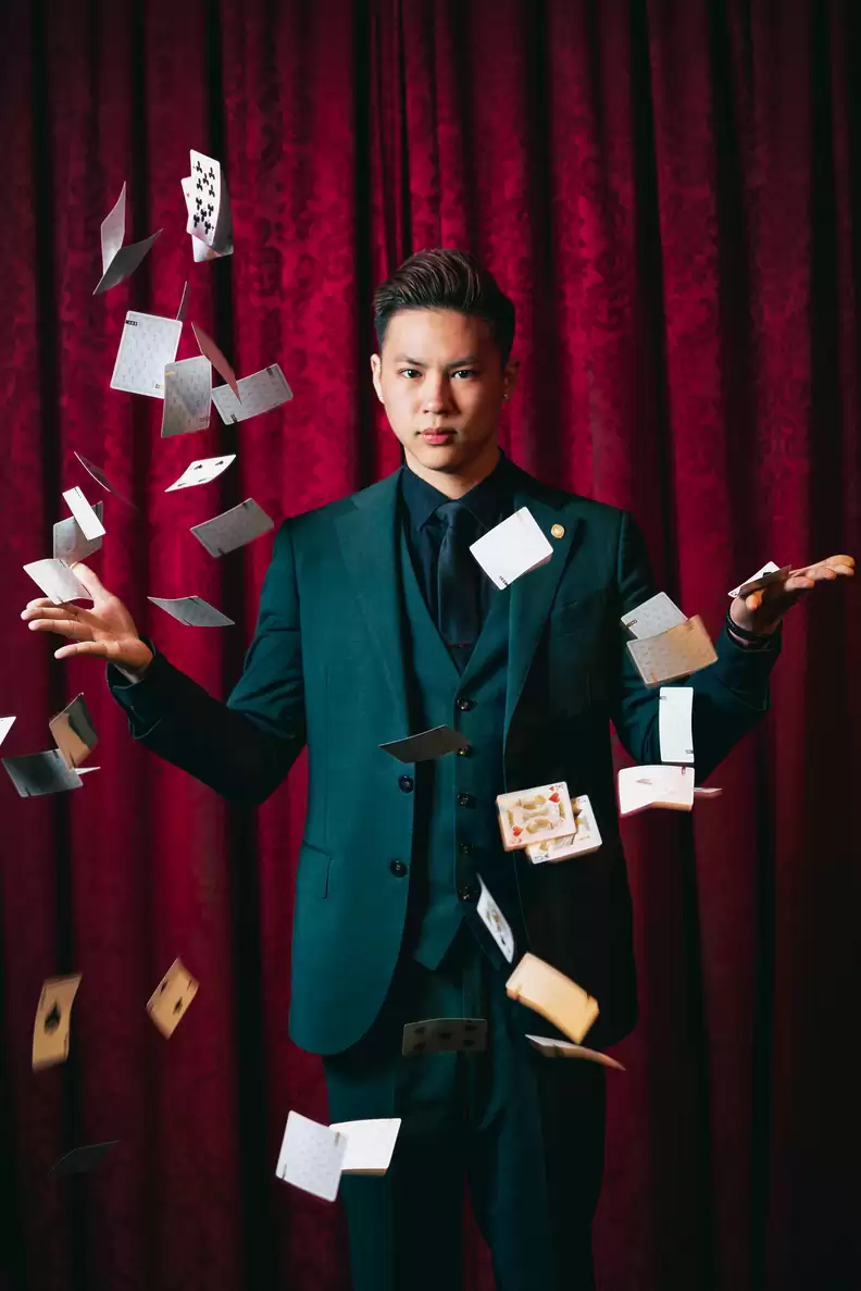 A magician in a formal suit performs card tricks with playing cards floating around him, set against a deep red curtain.