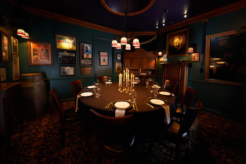 A dimly lit dining room with a black tablecloth, elegant table settings, and walls adorned with vintage posters and framed art.
