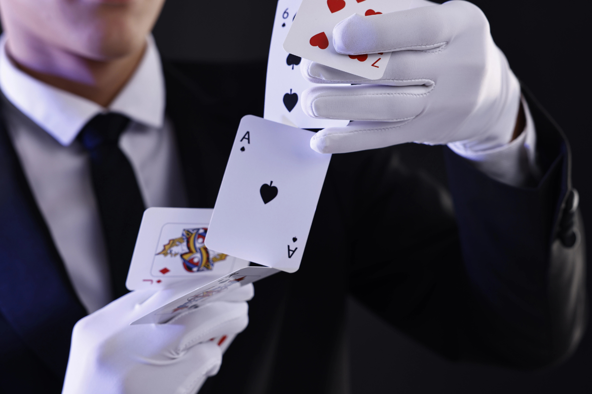 A magician in a black suit and white gloves reveals several playing cards, including an Ace of Spades, in a dramatic, illuminated pose.