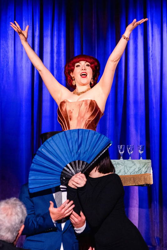 A performer with red hair raises hands on stage, while another person holds a blue fan, with tableware in the background.