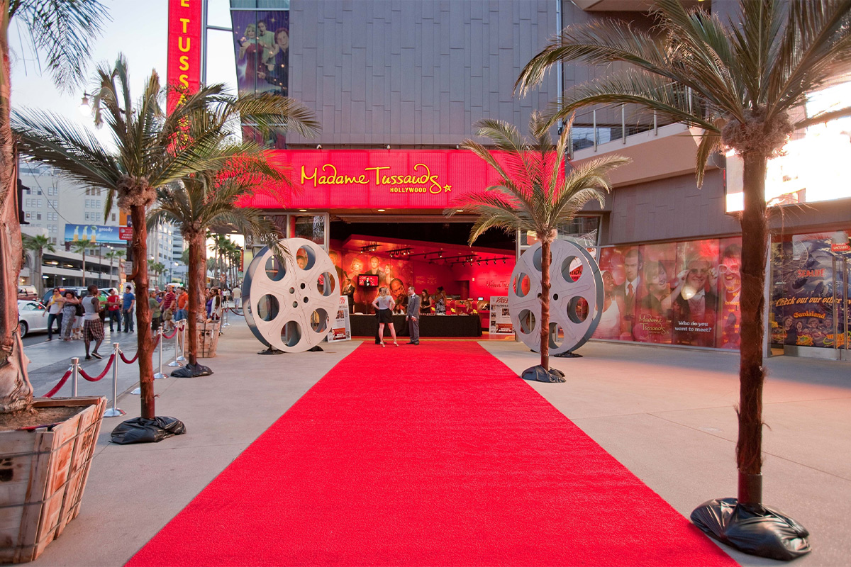 A vibrant red carpet leads to the entrance of Madame Tussauds Hollywood, flanked by palm trees and film reel decor.