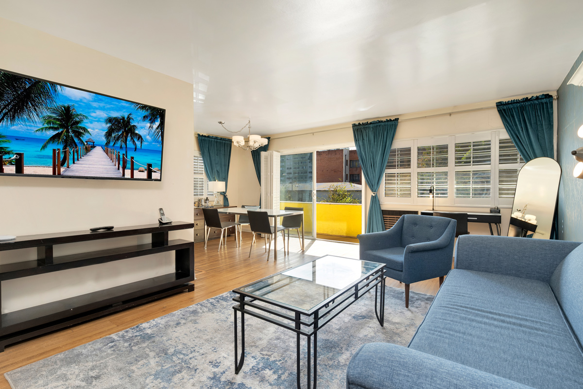 A bright, modern living room featuring a blue couch, a glass coffee table, and a large TV displaying a beach scene, with curtains and dining area visible.