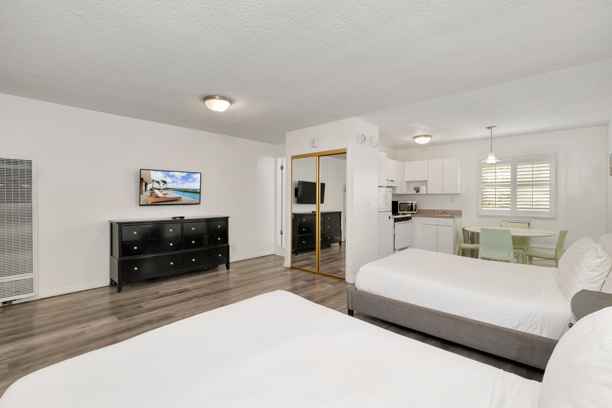 A bright and modern room featuring two beds, a television, and a kitchenette with dining area, adorned with light wooden flooring.