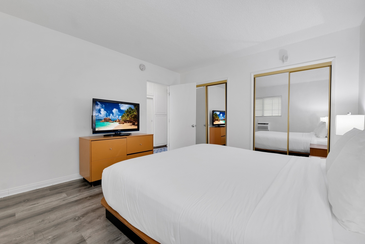 A modern bedroom featuring a large bed, a television on a dresser, and mirrored wardrobe doors, with light-colored walls and flooring.