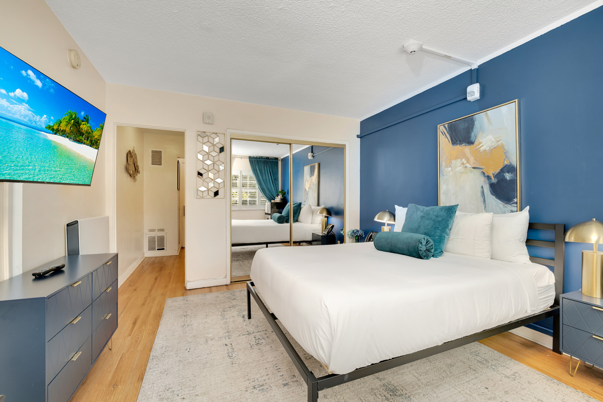 A modern bedroom with a blue accent wall, featuring a mirrored wardrobe, a king-size bed, and a TV displaying a beach scene.