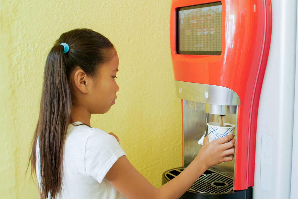 A young girl stands at a drink dispenser, filling a paper cup with a beverage, against a bright yellow wall.