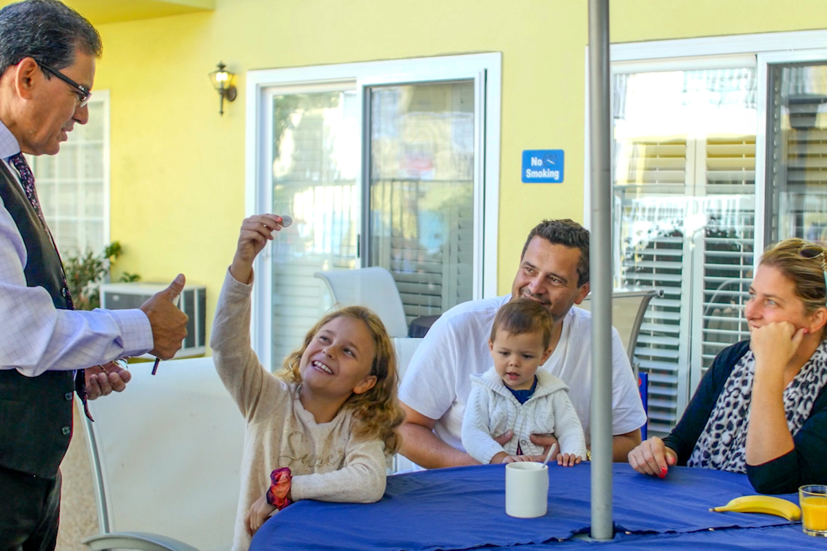 A magician entertains a family at a table outdoors, with a girl holding up a coin, while others watch and smile.