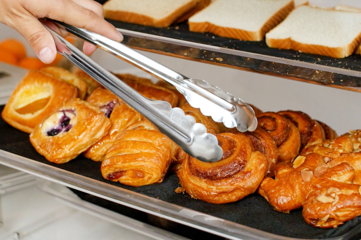 A selection of freshly baked pastries including croissants and breads, with a hand using tongs to serve.