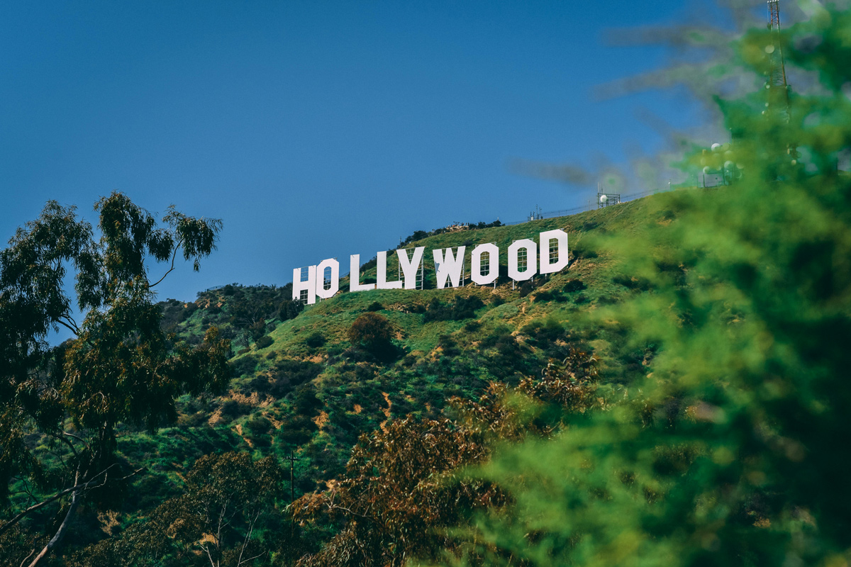 The iconic Hollywood sign is prominently displayed atop a green hill against a clear blue sky, surrounded by lush greenery.