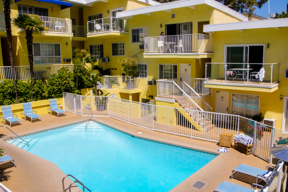 A sunny courtyard featuring a blue swimming pool surrounded by yellow buildings with balconies and palm trees.