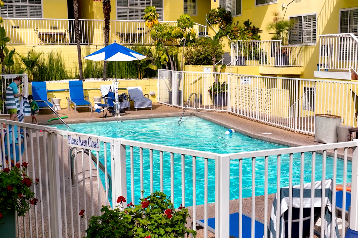 A bright outdoor pool area surrounded by lounge chairs, fences, and tropical plants, showcasing a sunny day atmosphere.