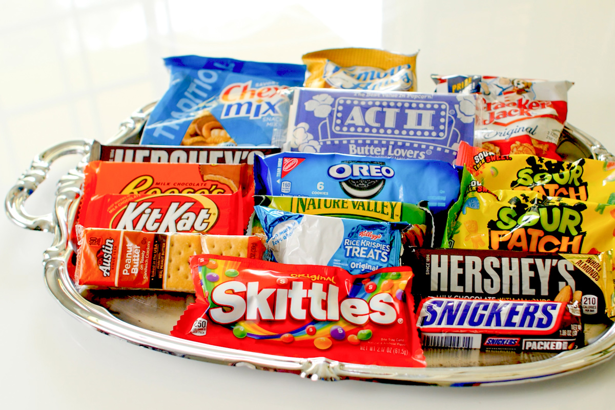 A variety of snacks arranged on a silver tray, including candies, cookies, and popcorn, showcasing colorful packaging and different brands.